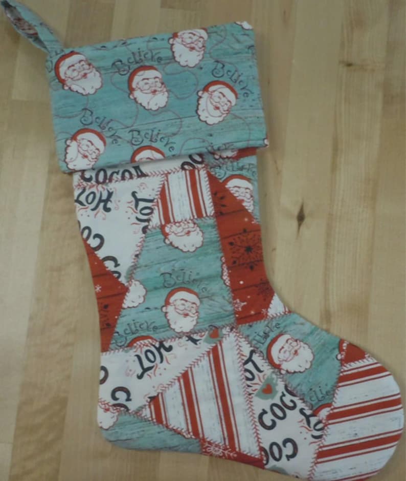 Op de afbeelding: Een kerstkous met een patchworkontwerp en afwisselende stoffen. De boord is lichtblauw met kerstmangezichten en het woord Believe. De kous zelf bevat rood-witte strepen, kerstmangezichten en de tekst Hot Cocoa in een decoratief patroon.