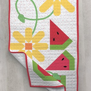 May include: A white quilt with a red border features a yellow flower with a green stem and leaves, and two red watermelon slices with green rinds. The background is white with small red dots.