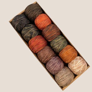 May include: A box of 12 balls of embroidery floss in shades of brown, orange, and green. The balls are arranged in a row, with the brown balls at the top and the green balls at the bottom.