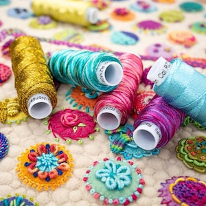 May include: A close-up of embroidery floss spools in various colors, including gold, teal, pink, blue, and purple. The spools are arranged on a white surface with colorful embroidered designs.
