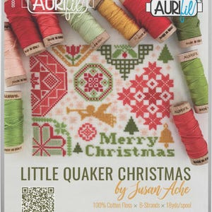 May include: A cross-stitch pattern with the text "Merry Christmas" and the title "Little Quaker Christmas." The design features red, green, and gold cross-stitch elements, including a Christmas tree, angel, and gift. Spools of Aurifil thread in various colors surround the pattern.