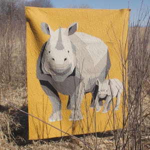 May include: A handmade quilt featuring a geometric design of a rhinoceros and its calf against a mustard yellow background. The rhino is primarily gray and white, with darker gray accents. The quilt is displayed outdoors.