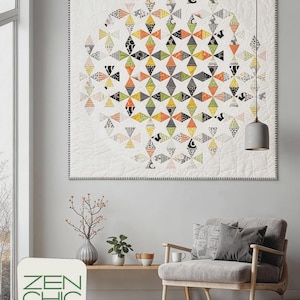 May include: A square quilt featuring a central circular pattern of small, multi-colored diamond shapes in orange, yellow, green, and black and white prints against a white background. The quilt has a thin, dark, patterned border.