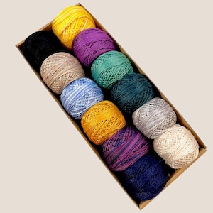 May include: A box of 12 colorful yarn balls in various shades of blue, green, purple, yellow, and beige. The yarn balls are arranged in a row in a cardboard box.