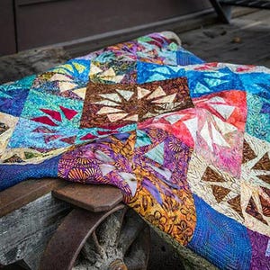 May include: A colorful quilt with geometric patterns, featuring shades of blue, purple, red, and brown. The quilt is draped over a rustic wooden cart wheel, with the website address "Quiltworx.com" visible in the upper left corner.