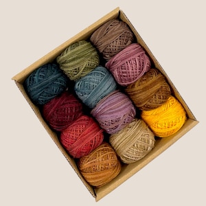 May include: A cardboard box filled with twelve colorful yarn balls. The yarn balls are in shades of blue, green, red, purple, brown, and yellow. The yarn is tightly wound and appears to be made of a soft material, perfect for knitting or crocheting projects.
