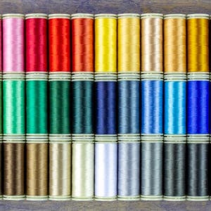 May include: A collection of 30 spools of thread in various colors, including purple, pink, red, orange, yellow, green, blue, brown, gray, and white. The spools are arranged in three rows of ten.