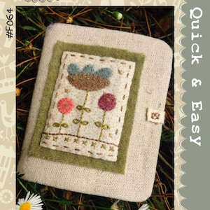May include: A beige needlebook with a floral design. The cover features a stitched rectangle with three flowers in pink, red, and blue. The text "Hatched and Patched" and "Little Flower Needlebook" are visible.