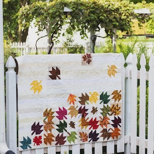 May include: A rectangular quilt featuring a cream-colored background with horizontal textured stripes. The quilt is decorated with numerous fabric maple leaf appliques in various autumn colors including red, orange, yellow, brown, and dark green.