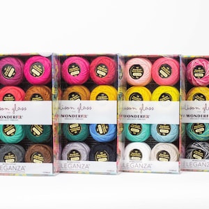 May include: Four boxes of embroidery floss, each containing 12 spools of thread. The boxes are arranged in a row from left to right. The first box contains pink, red, and orange thread. The second box contains yellow, green, and blue thread. The third box contains light blue, gray, and brown thread. The fourth box contains white, cream, and black thread. Each box has the text "Alison Glass for WONDERFIL" and "ELEGANZA" printed on it.