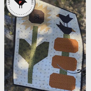 May include: A quilted wall hanging featuring a sunflower, pumpkins, and birds. The artwork is 14 inches by 20 inches. The design includes the text "PUMPKIN STACK" and "SCRAP BUSTER PATTERN". The artwork is displayed outdoors.