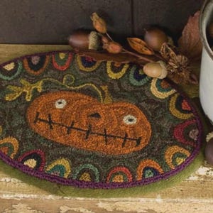 May include: Oval-shaped, hand-hooked rug featuring a smiling jack-o'-lantern design. The rug has a dark green background with a colorful border of rainbow-colored arches. A purple border frames the rug. Acorns and a metal container filled with acorns are in the background.