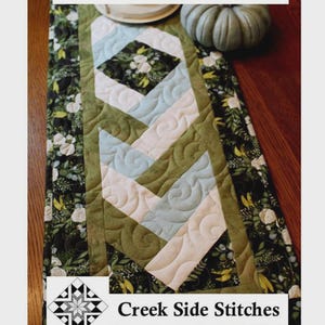 May include: Braided runner, 12.5 x 40 inches, with a quilted design. The runner features a central pattern of light blue and cream-colored diamonds and chevrons, framed by green fabric and floral borders. The text "Braided Runner" and "Creek Side Stitches" are visible.