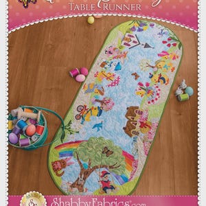 May include: An Easter Sunday table runner with a colorful design featuring bunnies, rainbows, and trees. The runner has a green border and the text "Easter Sunday Table Runner". A basket of spools and eggs is in the corner.