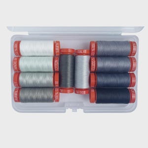 May include: A clear plastic case containing spools of Aurifil cotton thread in various shades of gray and white. The spools have red caps and are labeled with "100% Cotton" and "Made in Italy".