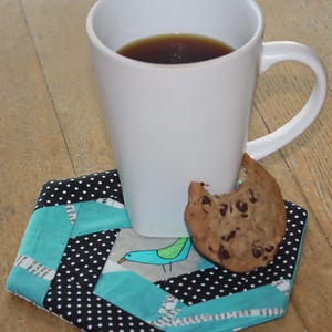 May include: A white ceramic mug filled with dark liquid sits on a hexagonal fabric coaster featuring a patchwork design of black fabric with white polka dots, solid teal fabric, and white fabric with black text, plus a chocolate chip cookie.
