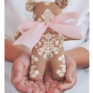 May include: A brown teddy bear with a pink bow, the bear has a white floral pattern on its body.