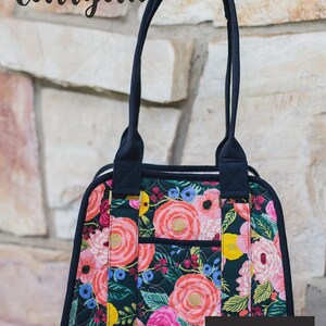 May include: A quilted tote bag featuring a dark background with a vibrant floral pattern of pink, red, and yellow flowers, accented by a solid magenta base. The bag has dark navy handles and trim, with the text Boundless Carryall and Knot + Thread design.