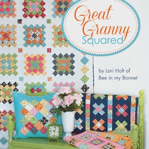 May include: A quilt pattern book titled Great Granny Squared by Lori Holt of Bee in my Bonnet, displayed with a colorful patchwork quilt, a turquoise pillow, a navy quilt, a pink quilt, a white vase with pink flowers, and a green alarm clock.