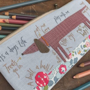 May include: A blue fabric pencil case with a brown felt dachshund dog applique. The dog has the text "Play with me" and "Rub my belly" stitched on it. The case also has the text "It's a dogs life", "Treats go in a ball", and "A ball, a ball!" stitched on it.