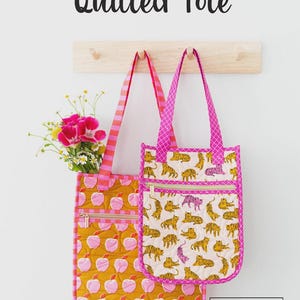 May include: Two quilted tote bags with pink accents hang on a wooden rack. One bag features a yellow apple pattern, the other a tiger print. Both have zippered pockets and pink straps. The text "Everyday Quilted Tote" is at the top.
