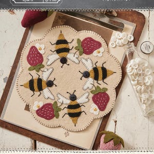 May include: A round, beige felt mat with a scalloped edge, featuring embroidered bees and strawberries. The bees have black and yellow stripes, and the strawberries are red. The text "BEES AND STRAWBERRIES ALL AROUND" is visible. The mat is 8.5 inches in diameter.