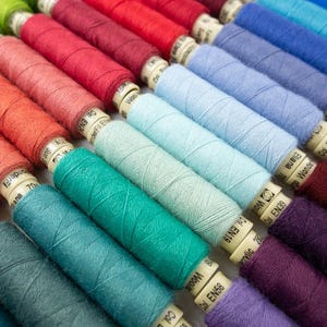 May include: A collection of spools of thread in various colors, including green, red, blue, teal, purple, and brown. The spools are arranged in a rainbow pattern and have labels with numbers and letters.
