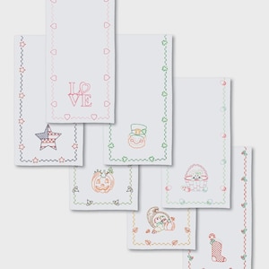 May include: White embroidered decorative towels with various holiday and seasonal designs. The designs include a patriotic star, a snowman, a pumpkin, a bunny, a cornucopia, a stocking, and the word "LOVE". Each towel has a decorative border.