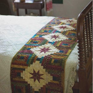 May include: A rectangular quilted bed runner featuring a repeating pattern of cream-colored stars centered within multi-colored log cabin blocks. The runner displays a palette of deep red, forest green, navy blue, and gold fabric strips with intricate stitching.