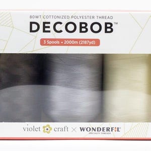 May include: A box of DECOBOB cottonized polyester thread with three spools visible through a rectangular window. The spools are in shades of gray and cream. The box has the text "DECOBOB" and "3 Spools - 2000m (2187yd)". The brand names "violet craft" and "WONDERFIL" are also visible.