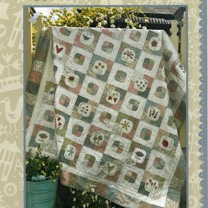 May include: A quilt pattern featuring a patchwork design with a variety of floral and geometric motifs. The quilt is displayed against a white picket fence and green foliage. The pattern is titled "Market Garden Quilt" and is from the "Hatched and Patched" collection.