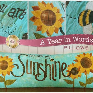 May include: A rectangular turquoise pillow with the words "You are my Sunshine" and two sunflowers. The pillow also features a small bee and the words "You are" at the top. The words "A Year in Words" and "Pillows" are also visible.