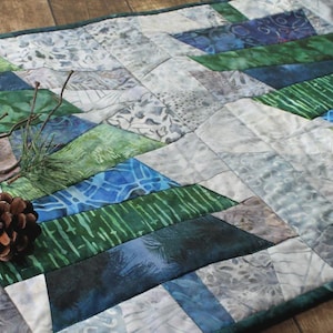 May include: A quilted table runner with a geometric design in shades of blue, green, and gray. The runner is decorated with pine cones and sprigs of pine needles. The runner is approximately 40 inches long.