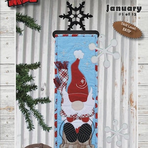 May include: A fabric wall hanging featuring a gnome design, titled "Blizzard Billy." The design includes a red hat, scarf, and boots, set against a blue background with snowflakes. The text reads "January #1 of 12" and is 6" x 20".