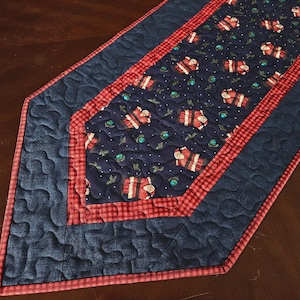 May include: A quilted table runner featuring a central navy blue panel with a repeating Santa Claus and holly print, bordered by a red and black checkered fabric and a solid navy blue outer section with a meandering stippled stitching pattern.
