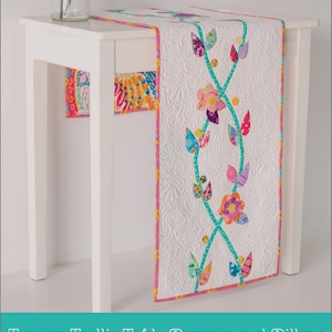 May include: White table runner with a floral trellis design. The runner features colorful appliqued flowers and leaves on a white quilted background. The edges are trimmed with a pink and yellow border. The text "Tuscan Trellis Table Runner and Pillow" is at the bottom.