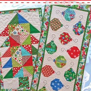 May include: Two Christmas-themed table runners. One features a quilted Christmas tree design with red, green, and blue triangles. The other has quilted ornament designs in red, green, and blue with the text "Joy Ho!" and "Material Girlfriends" in the corner.
