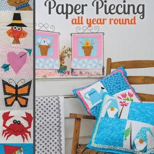 May include: A book titled "Paper Piecing all year round" by Mary Ann Hertel, with colorful patchwork designs. The image also shows decorative pillows and fabric samples, all featuring whimsical, sewn designs. The book cover and pillows showcase various patterns.
