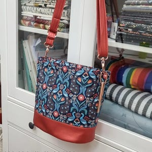 May include: An oval bag with a vibrant peacock and floral pattern on a navy blue background. The bag has a red base and matching straps. The bag measures 10" wide, 9" high, and 6.5" deep. The text "The Awesome Oval Bag" is at the top.