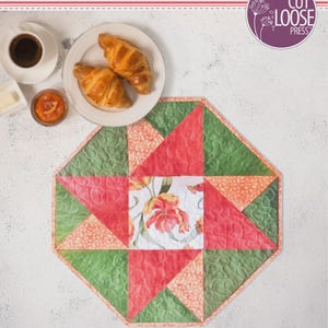 May include: A quilted octagonal table topper titled Splendid Spinner Topper designed by Cathey Laird. The fabric features a central floral square surrounded by alternating red, green, and orange patterned triangles. It measures 19.5 by 19.5 inches.