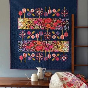 May include: A navy blue quilt with floral patterns and the text "Gathering Wildflowers." The quilt features rows of colorful flowers and geometric shapes. The quilt is displayed on a wall above a wooden table.