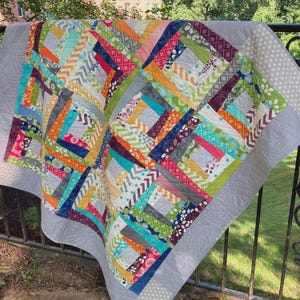 May include: A patchwork quilt featuring a log cabin block pattern with a variety of colorful fabric strips in shades of orange, pink, green, blue, and yellow. The quilt is bordered by a solid light gray fabric with a subtle quilted diamond pattern.