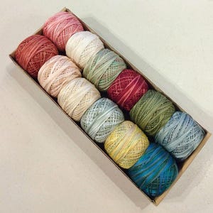 May include: A set of 10 balls of yarn in various shades of green, blue, red, and beige. The yarn is wound into balls and arranged in a row in a cardboard box.
