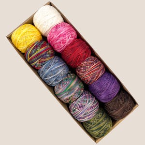 May include: A rectangular cardboard box filled with twelve spools of colorful embroidery thread. The spools are arranged diagonally and feature a variety of colors, including yellow, pink, red, blue, green, purple, and brown. The thread is tightly wound.