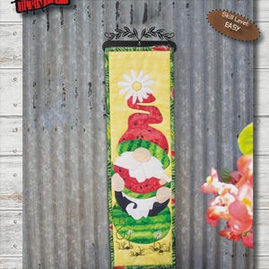 May include: A decorative wall hanging featuring a gnome design with a watermelon theme. The gnome is wearing a watermelon hat and holding a watermelon. The banner is approximately 6 inches wide and 20 inches long.