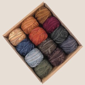 May include: A cardboard box filled with twelve skeins of yarn in various colors. The yarn colors include shades of orange, brown, blue, green, and gray. The yarn is tightly wound into small balls, perfect for knitting or crocheting projects.