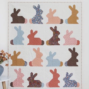May include: A white quilt with a repeating pattern of bunny silhouettes in various colors and patterns. The word "Bunnies" is at the top. The text "A fat quarter quilt in 3 sizes" is at the bottom. The quilt is displayed on a white wall.