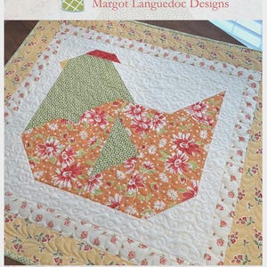 May include: A quilted nesting table topper with a bird design. The bird is made of orange floral fabric and green patterned fabric, set against a white background. The topper measures 28 inches by 30 inches.