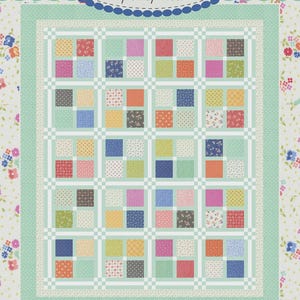 May include: A quilt pattern design featuring a grid of colorful squares in various patterns and colors, including pink, blue, orange, and green. The design is framed by a light green border and floral patterned fabric. Text reads "Coriander Quilts by Corey Yoder" and "#217 Like A Charm."