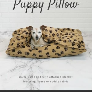 May include: A tan dog bed with a paw print pattern and attached blanket. The words "Puppy Pillow" are at the top. A small dog is nestled inside the bed. The text "snuggly dog bed with attached blanket featuring fleece or cuddle fabric" is below the bed.